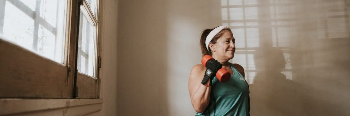 Senior woman in a brightly lit room exercises with dumbbells, promoting fitness at home.